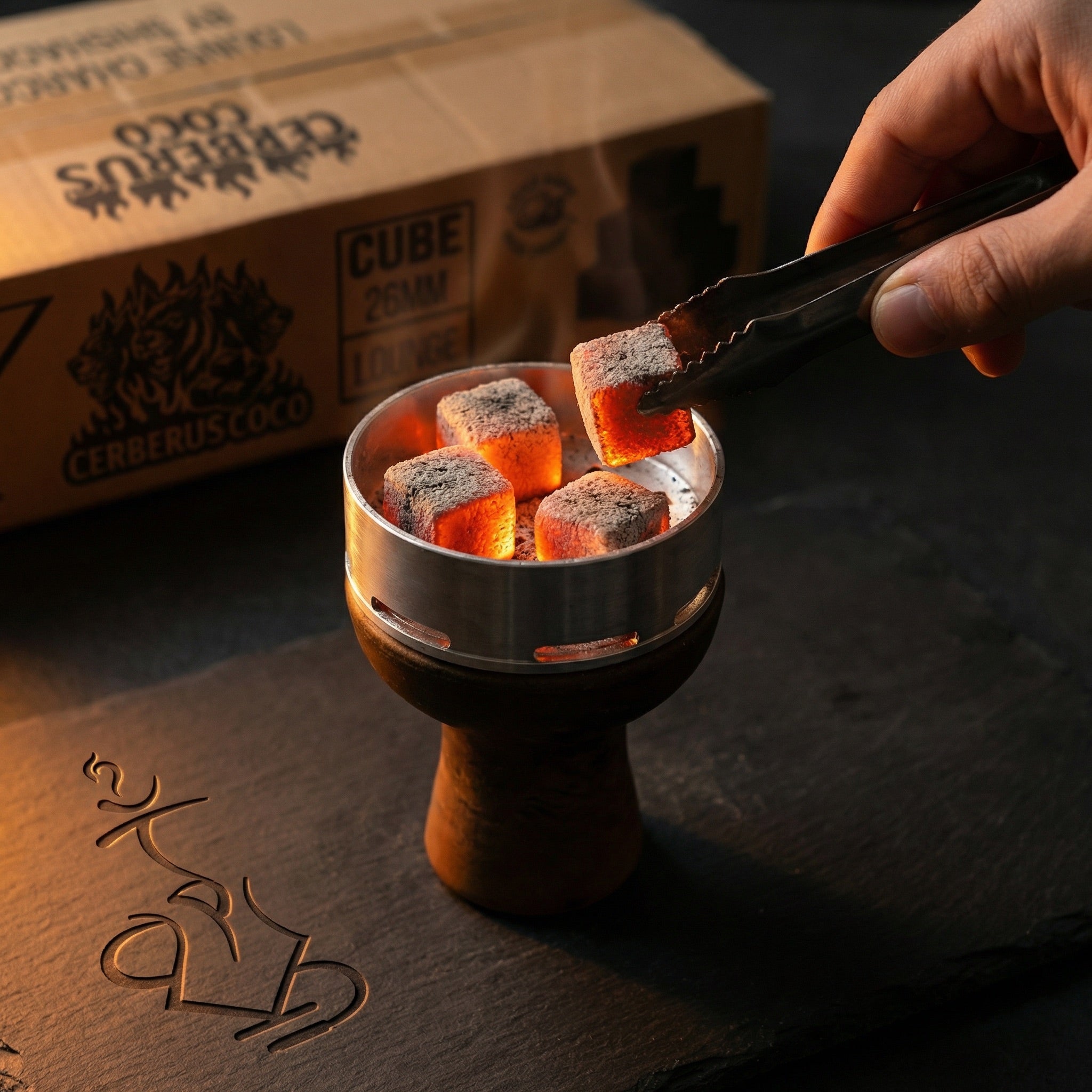  Glowing Cerberus Coco 26mm coconut shisha charcoal cubes inside a heat management device on a hookah bowl, hand placing a coal with tongs during a session.
