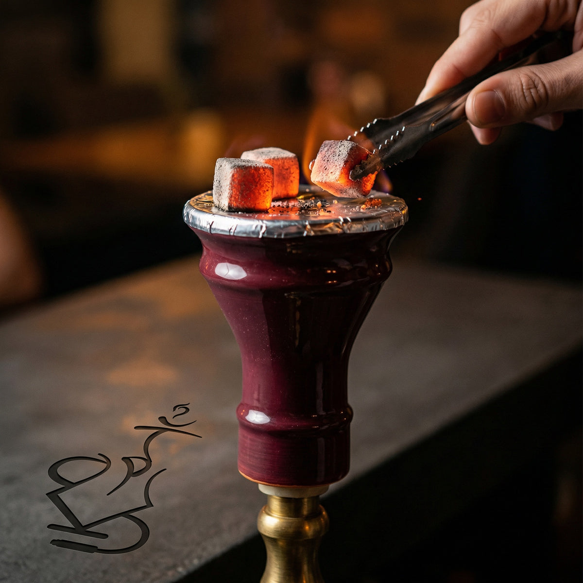 King Buzz Traditional Egyptian Bowl foiled with glowing coals on a hookah, a hand rotating charcoal with tongs in a warm dimly lit session setting., image 2 of 7