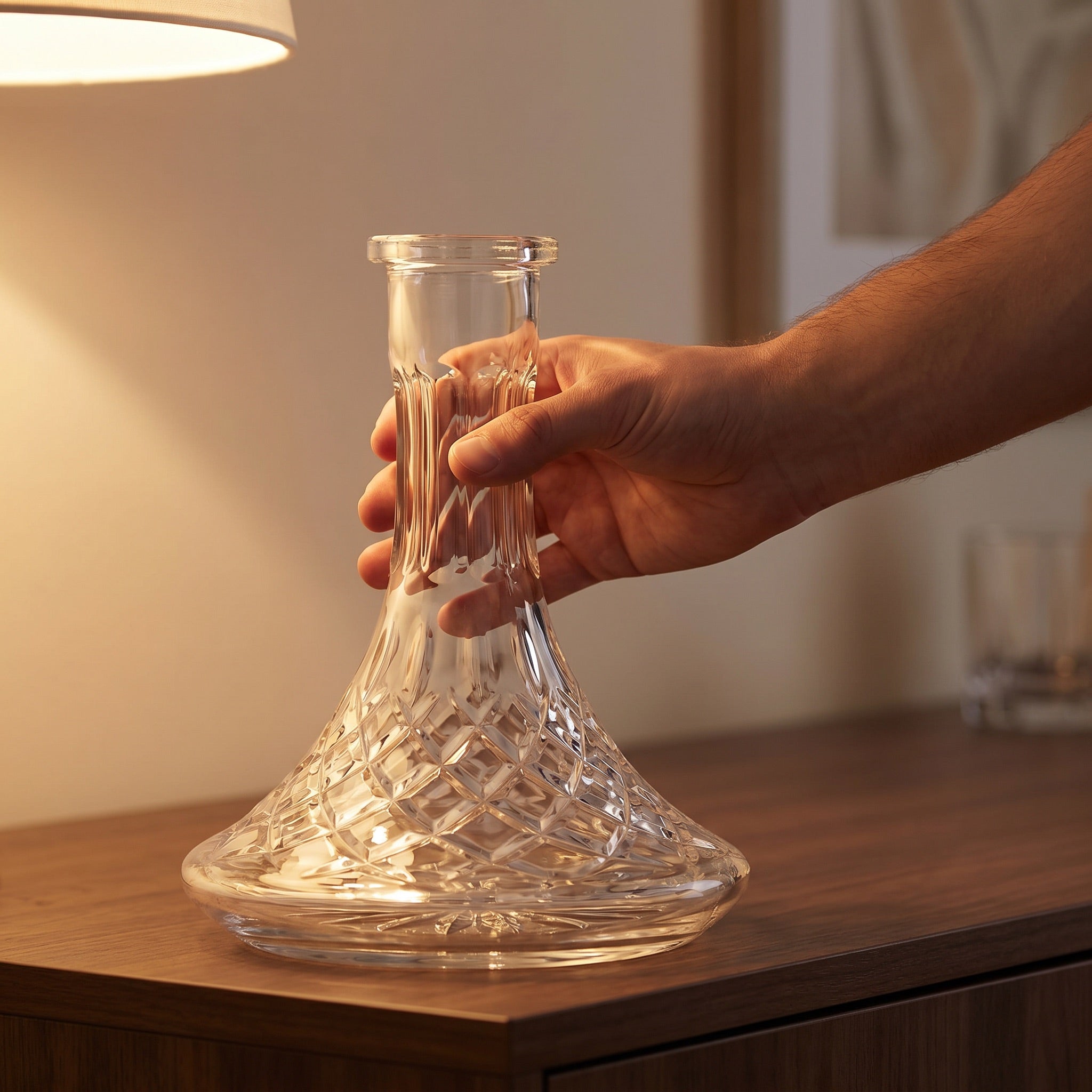 Hand lifting Lismore crystal-cut hookah base from dark wood sideboard in warm lamplit home setting