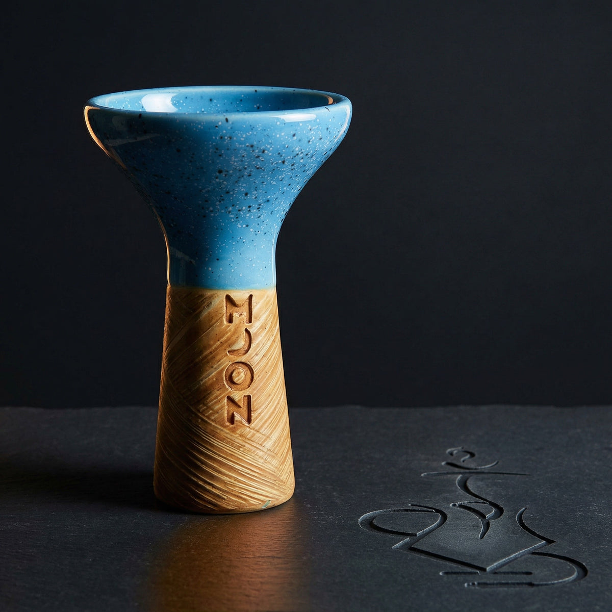 Moon Phunnel blue speckled glazed clay shisha bowl shot in dramatic dark studio lighting on slate, the glaze and hand-stamped MOON wordmark glowing under a single warm key light., image 4 of 11