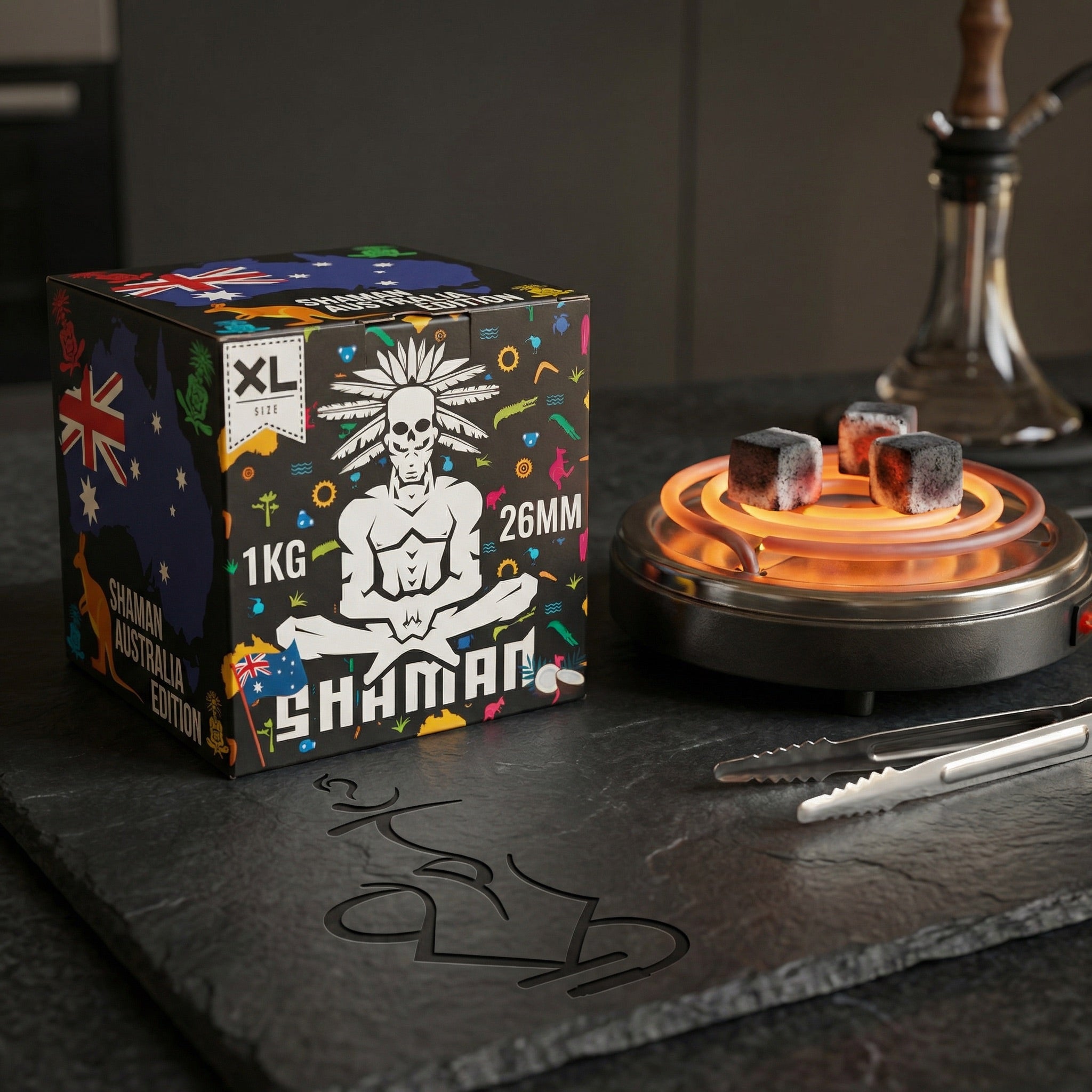 Shaman 26mm coconut charcoal 1kg box on a dark slate countertop beside an electric burner with glowing coals, tongs, and a hookah in the background.