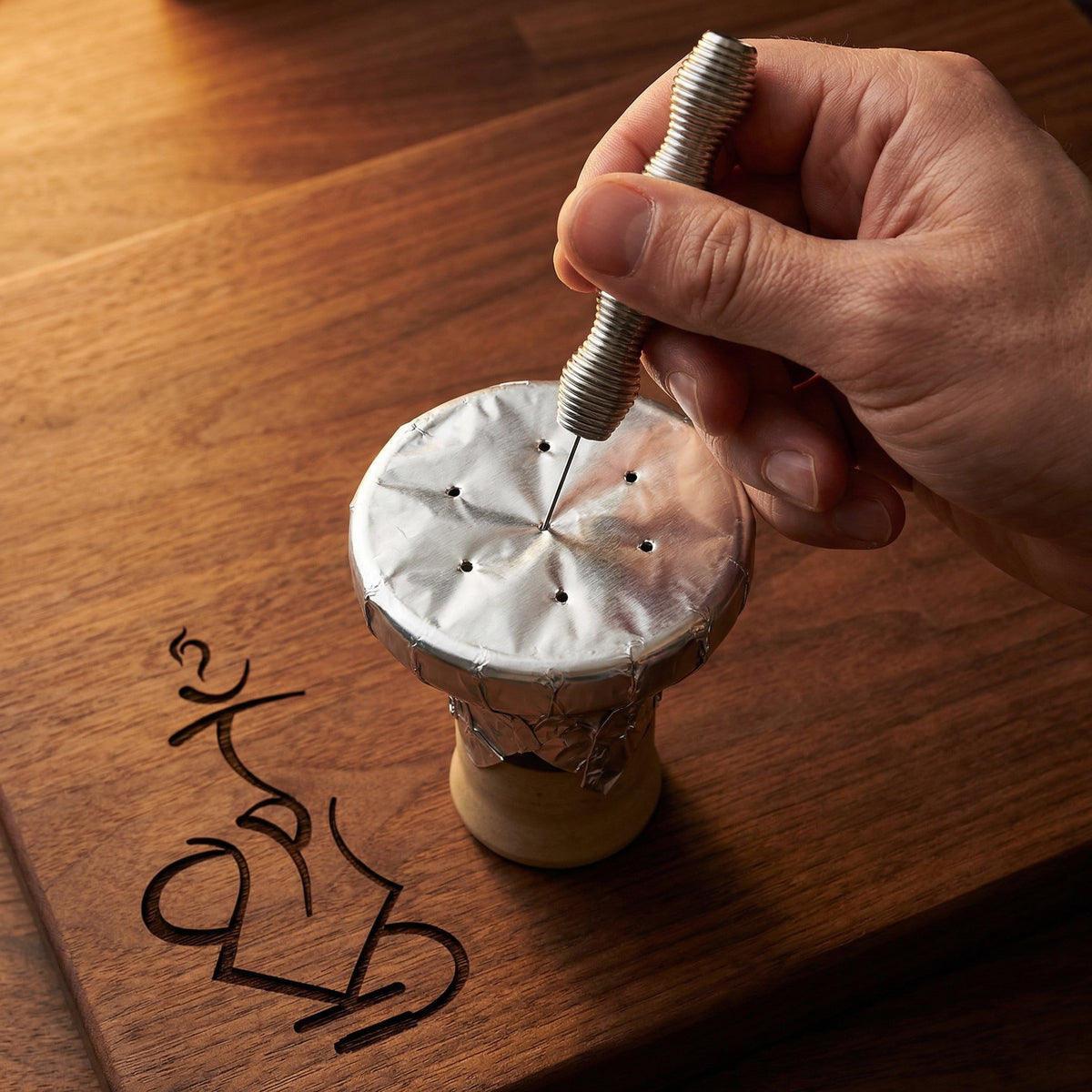A hand using the Smoothpoke ribbed stainless steel foil poker to punch airflow holes through taut foil stretched over a ceramic shisha bowl on a warm walnut prep surface., image 8 of 12